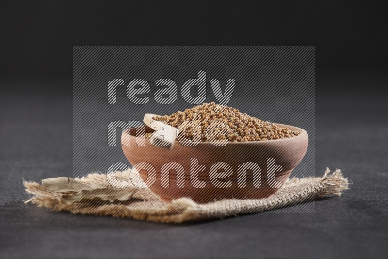 A wooden bowl and spoon full of mustard seeds on a piece of burlap on a black flooring
