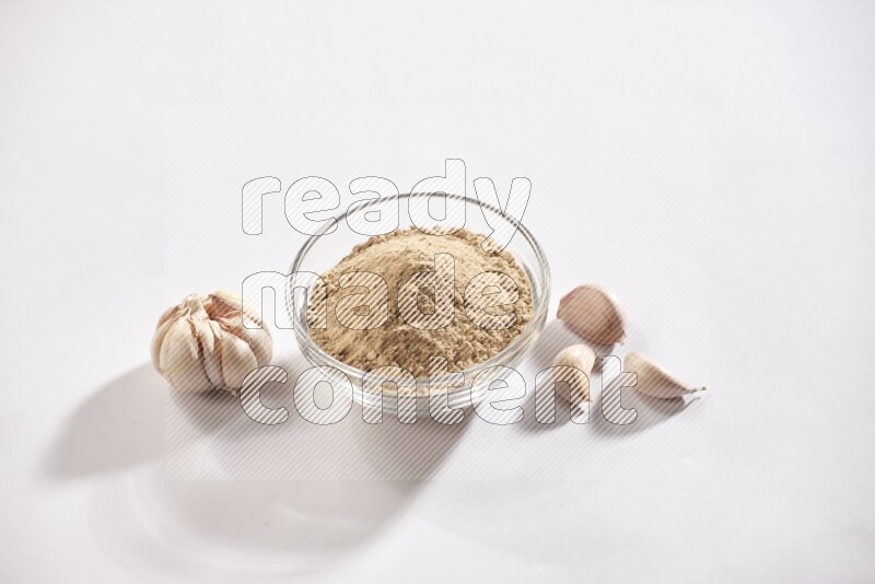 A glass bowl full of garlic powder with garlic bulb and some cloves beside it on a white flooring