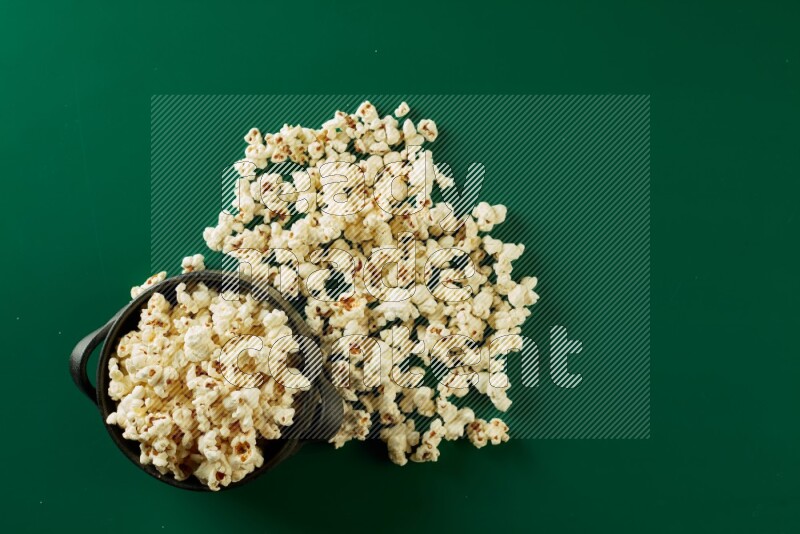 A black ceramic bowl full of popcorn with popcorn beside it on a green background in a top view shot