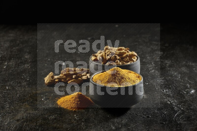 2 pottery black bowls, one full of turmeric powder and the other full of dried turmeric whole fingers on a textured black flooring
