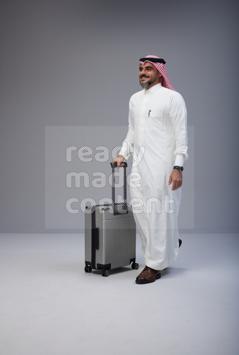 Saudi man wearing Thob and red Shomag standing holding Travel bag on Gray background