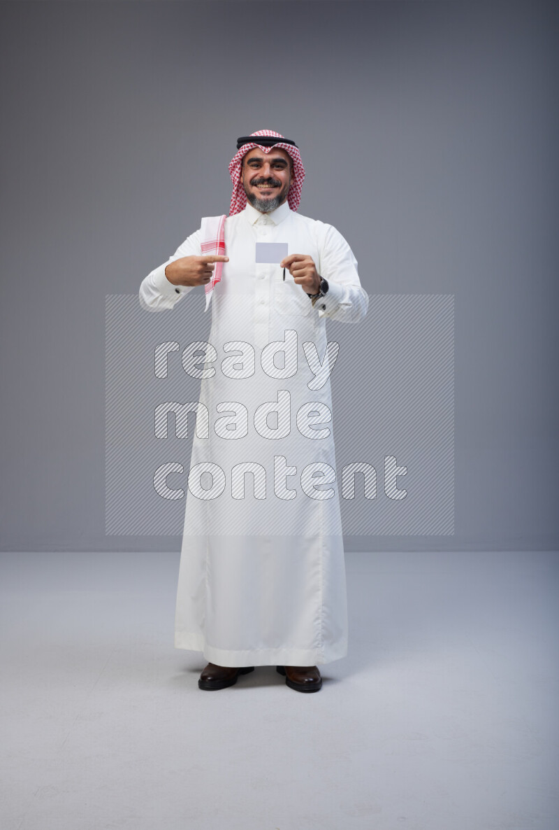 Saudi man Wearing Thob and red Shomag standing holding ATM card on Gray background