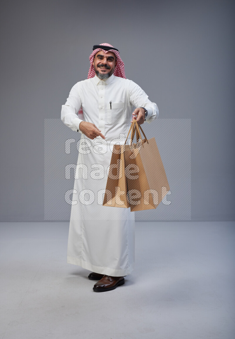 Saudi man Wearing Thob and red Shomag standing holding shopping bag on Gray background