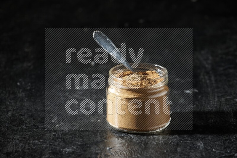 A glass jar and a metal spoon full of allspice powder on a textured black flooring