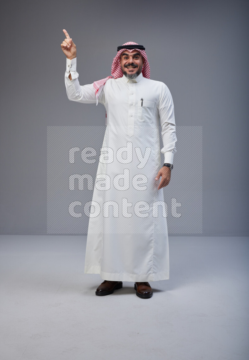 Saudi man Wearing Thob and red white Shomag standing interacting with the camera on Gray background