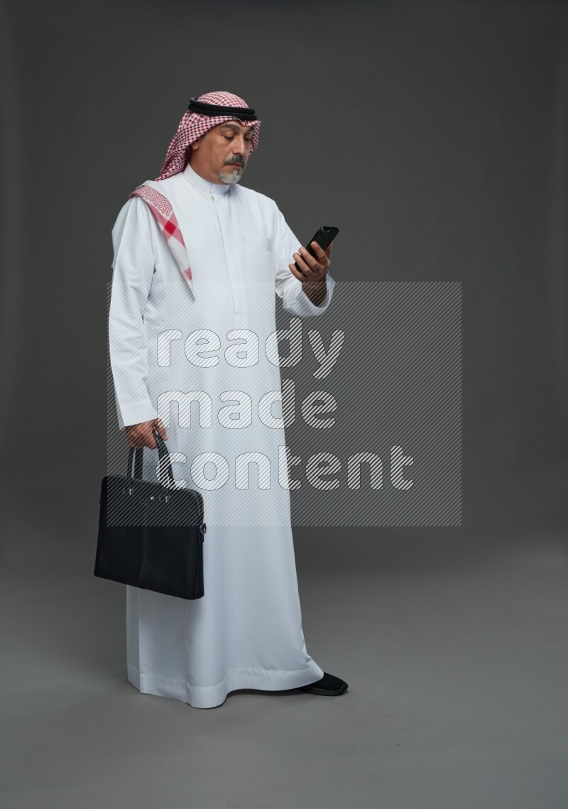 Saudi man with shomag Standing holding bag and phone on gray background