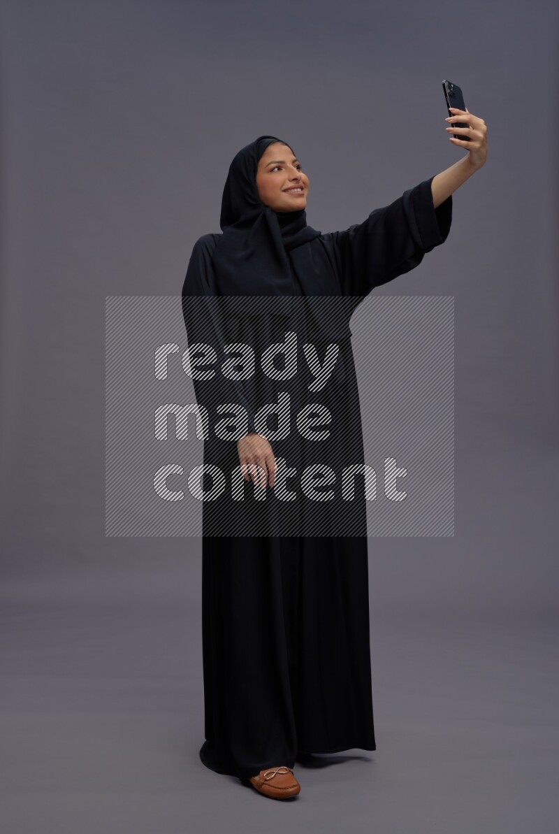 Saudi woman wearing Abaya standing taking selfie on gray background