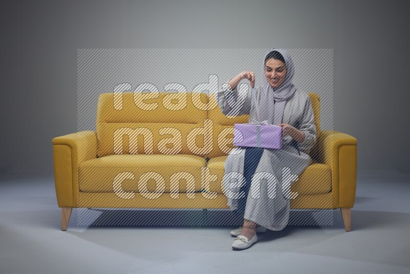 A Saudi woman wearing a light gray Abaya and head scarf sitting on a yellow sofa holding a gift box beside multi color gift boxes eye level on a grey background