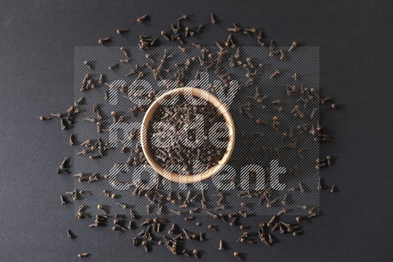 A wooden bowl full of cloves with spreaded whole cloves on a black flooring