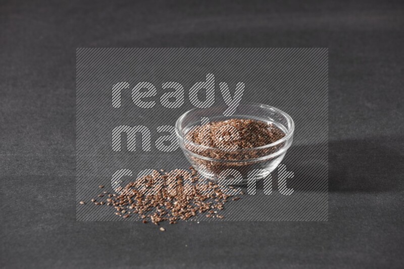 A glass bowl full of flaxseeds surrounded by the seeds on a black flooring