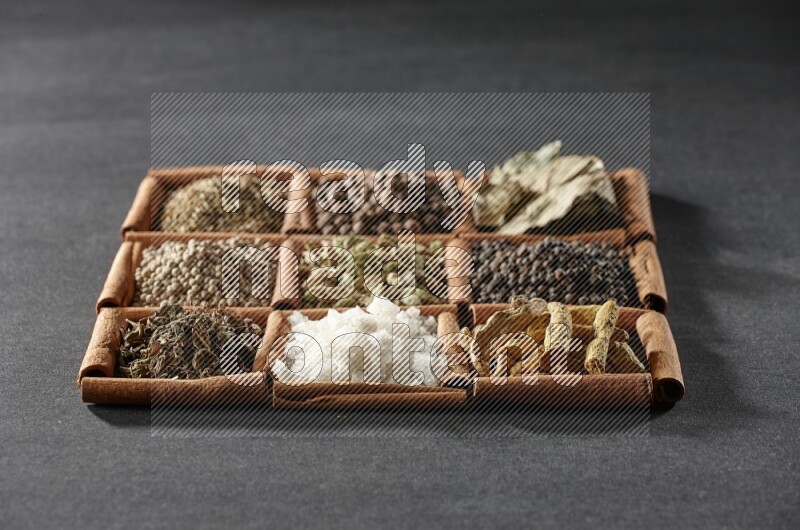 9 squares of cinnamon sticks full of cardamom in the middle surrounded by turmeric fingers, black pepper, bay laurel leaves, allspice, cumin, white pepper, dried basil and salt on black flooring