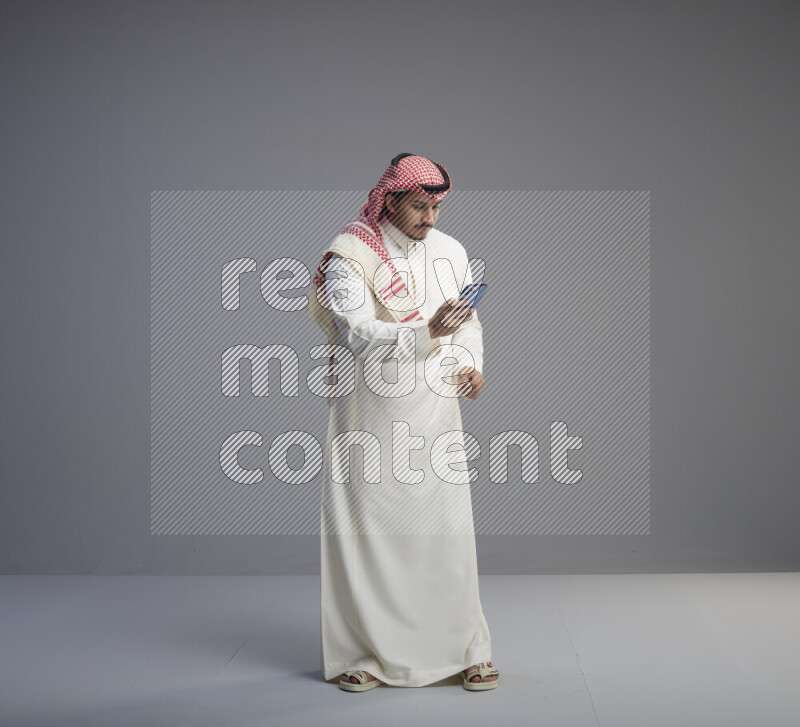 A  Saudi man standing wearing thob and red shomag texting on phone on gray background