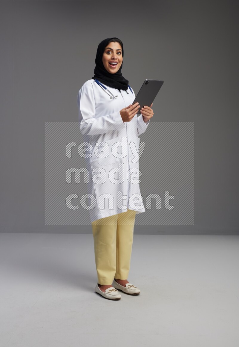 Saudi woman wearing lab coat with stethoscope standing working on tablet on Gray background