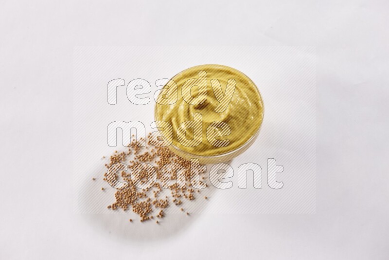 A glass bowl full of mustard paste with mustard seeds underneath on white flooring