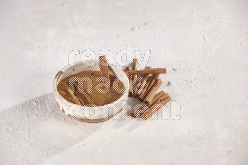 Ceramic bowl full of cinnamon powder with cinnamon sticks on the side on white background
