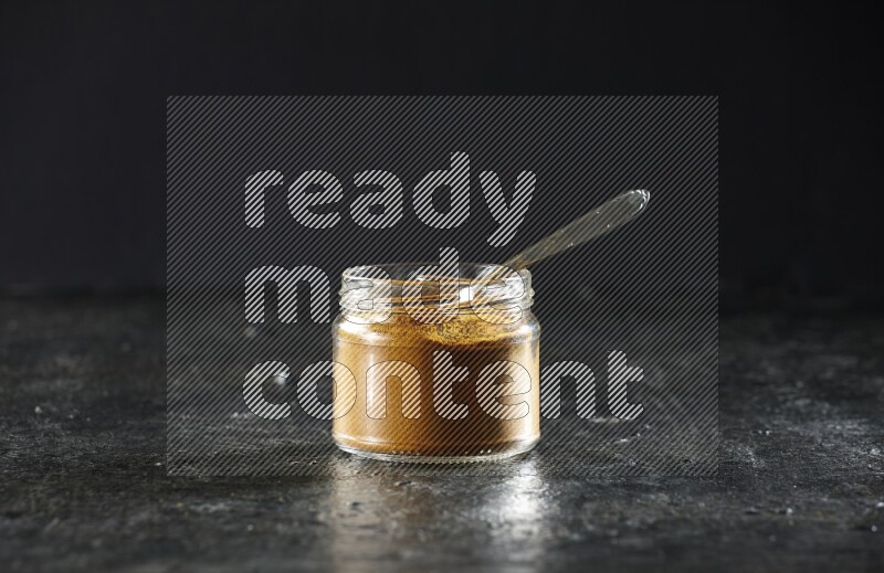 A glass jar and a metal spoon full of turmeric powder on a textured black flooring