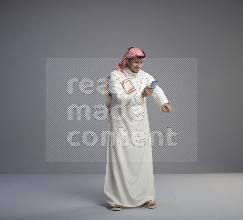 A  Saudi man standing wearing thob and red shomag texting on phone on gray background
