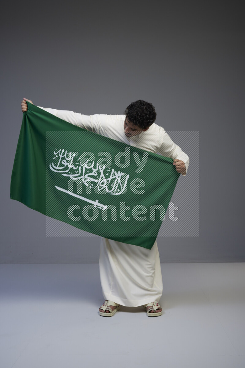 A Saudi man standing wearing thob holding big Saudi flag on gray background