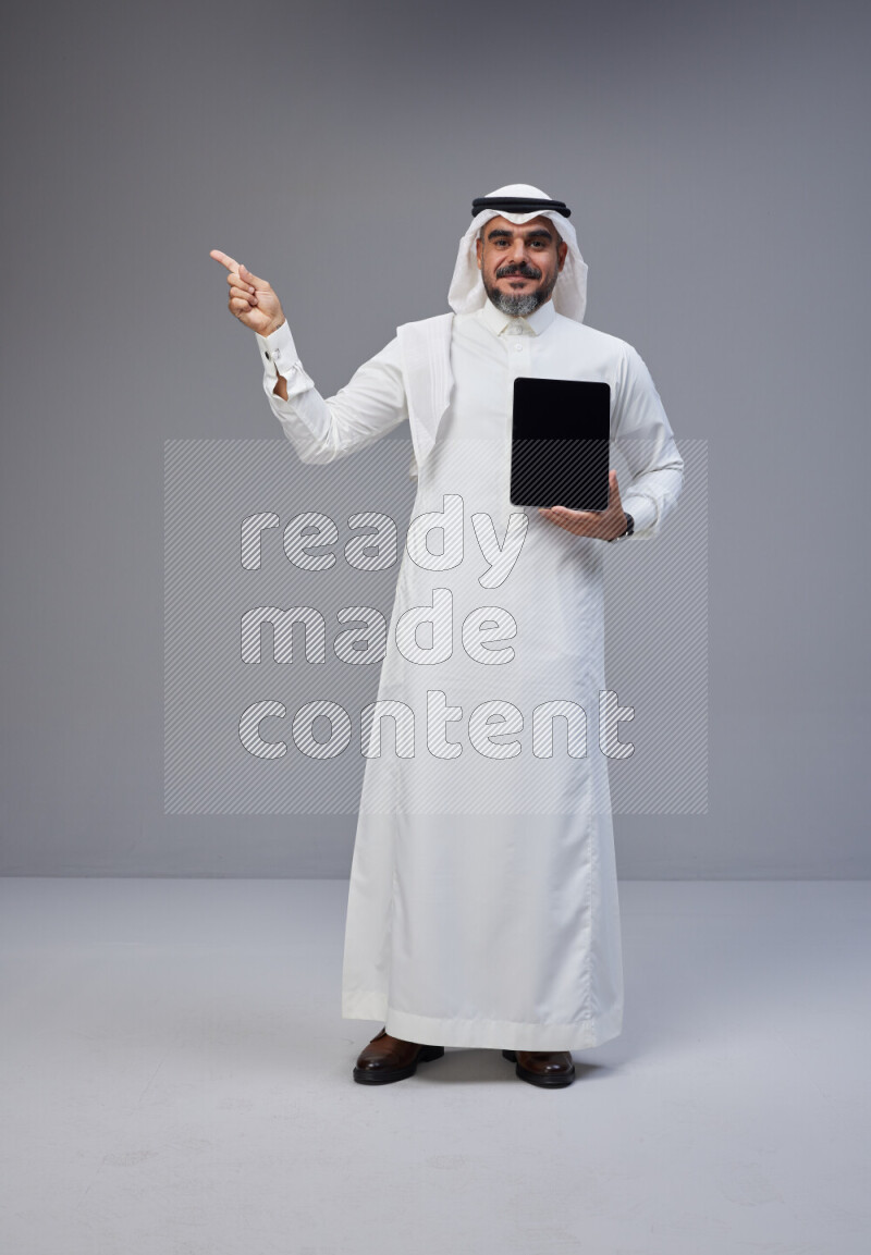 Saudi man Wearing Thob and white Shomag standing showing tablet to camera on Gray background