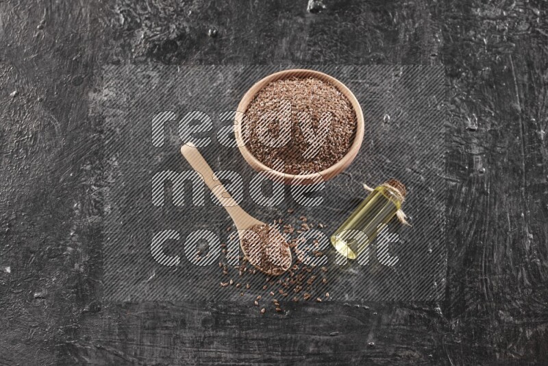 A wooden bowl and spoon full of flaxseeds and a glass bottle of flaxseeds oil on a textured black flooring