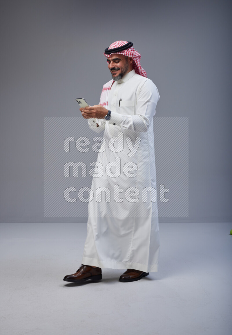 Saudi man Wearing Thob and red Shomag standing texting on phone on Gray background
